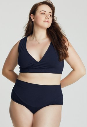 Donna curvy con capelli castano lunghi che indossa un costume da bagno blu navy a due pezzi, in piedi con le mani dietro la schiena, guarda verso destra.