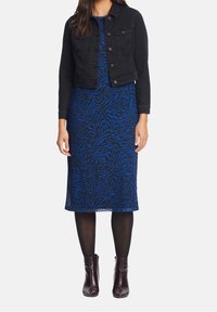 Žena v modro-černě vzorovaných šatech ke kolenům, černé krátké denimové bundě, černých punčochách a tmavohnědých kotníkových botách.