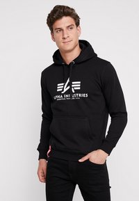 Zwarte katoenen hoodie met een voorkantzak, capuchon met trekkoord en witte logo-tekst. Ribgebreide mouwen en onderrand, met een klein rood label aan de zijkant.