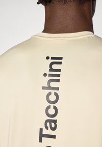 Ανοιχτό μπεζ μπλουζάκι με λεία υφή, που διαθέτει μία μεγάλη μαύρη εκτύπωση της λέξης "Tacchini" κάθετα κατά μήκος της πλάτης.