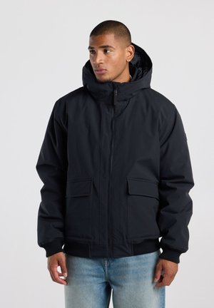 Man met een zwarte winterjas met capuchon en voorzakken en een blauwe spijkerbroek, die zijwaarts kijkt tegen een effen achtergrond.