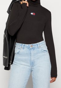 Person som bär en svart långärmad polotröja med Tommy Jeans-logga, ljusblå högmidjade jeans och håller en svart läderjacka över axeln.