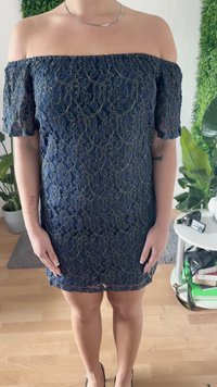 Mulher a usar um vestido de renda azul marinho, ombros à vista, de pé dentro de casa junto a plantas e uma mesa redonda com câmaras e livros.