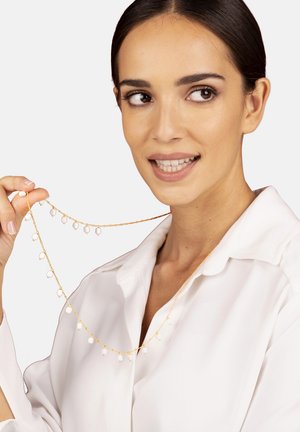 Goldkette mit kleinen weißen Perlen und runden Akzenten, zwischen den Fingern drapiert, vor einem neutralen Hintergrund.