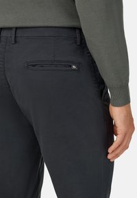 Tmavomodré chinos s hladkou textúrou majú zadnú vrecko so zipsom a malým kovovým logom. Nohavice sú kombinované so šedým sveterom.