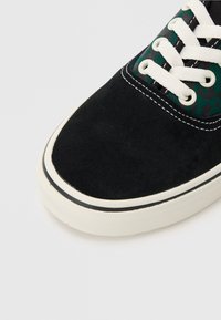 Svarta mockasneakers med vita snören, med ett grönt mönstrat tygpanel nära tungan och en vit gummisula med svarta detaljer.