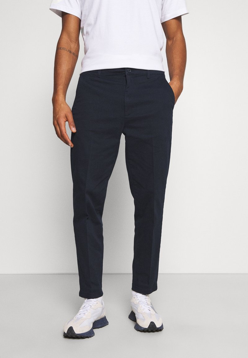 Homme portant un pantalon slim bleu marine, des baskets blanches et un t-shirt blanc, debout, une main dans la poche, devant un fond uni.