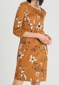Šaty s květinovým vzorem v teplé hořčicové barvě, s tříčtvrtečními rukávy a přiléhavým střihem, zdobené pestrobarevnými květy a listy.