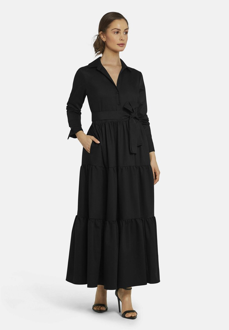 Schwarzes Maxikleid mit Kragen, langen Ärmeln und Taillengürtel. Mit gestuften Rüschendetails am Saum und seitlichen Taschen. Aus glattem Stoff.