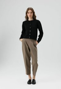 Cardigan noir boutonné avec un col rond, associé à un pantalon taille haute marron clair. Le modèle se tient les mains dans les poches, portant des mocassins noirs.