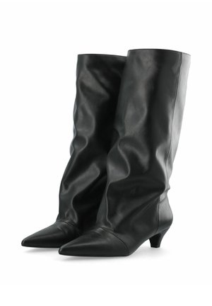 Paire de bottes en cuir noires montantes jusqu'au genou, à bouts pointus et petits talons kitten sur fond blanc.