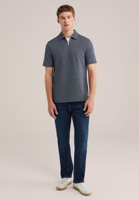 Tengerészkék, rövid ujjas póló zipzáras gallérral, sötétkék farmernadrággal és fehér sportcipővel kombinálva, semleges háttér előtt.