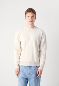 Cremfarbener, gerippter Strickpullover mit rundem Halsausschnitt und langen Ärmeln, kombiniert mit hellblauen Jeans. Glatte Textur, figurbetontes Design.