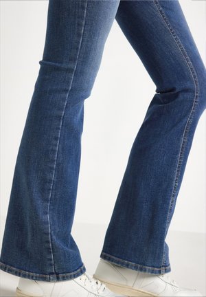 Jambes portant un jean évasé bleu foncé et des baskets blanches à lacets sur un fond clair uni.