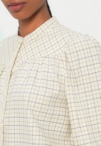 Beige bluse med et rutenetmønster i lys brun og sort. Har en kravehals og samlede detaljer omkring skuldrene.