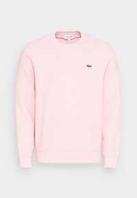 Felpa rosa chiaro in tessuto morbido, con scollo rotondo, polsini a coste e logo Lacoste verde sul petto a sinistra.