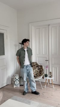 Kortärmad olivgrön skjorta över en vit t-shirt, ljusblå jeans och mörka skor, står i ett rum med vita dörrar och möbler.