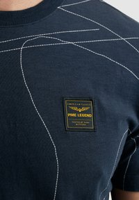 Navy blue t-shirt with white contrasting stitching lines, featuring a square patch label with yellow text and eagle design on the front.