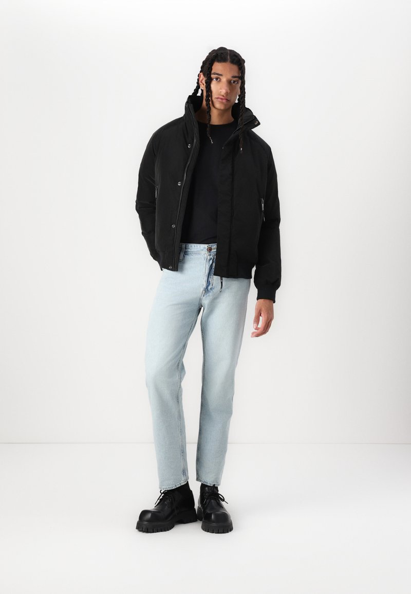 Chaqueta negra acolchada con cuello alto, botones frontales y bolsillos laterales; combinada con jeans rectos de mezclilla azul claros y botas chunky negras.