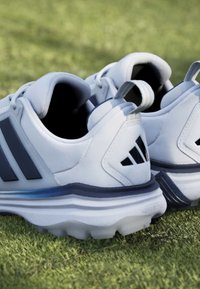Λευκά και γκρι αθλητικά παπούτσια με καρφιά που ακουμπούν σε πράσινο γρασίδι, δείχνοντας πίσω και πλευρικές λεπτομέρειες και των δύο παπουτσιών.