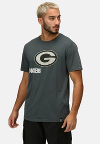 Šedé bavlněné tričko s výrazným kulatým logem s stylizovaným "G" a textem "PACKERS" pod ním. Klasický design s kulatým výstřihem.