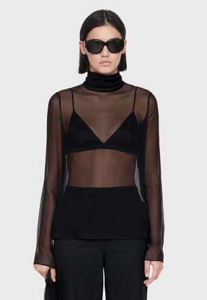 Woman wearing large black sunglasses, a sheer black long-sleeve top over a black bralette, and black pants against a plain background.