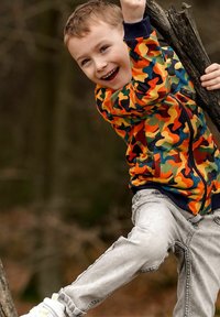 Młody chłopiec uśmiechający się i wspinający na gałąź drzewa na świeżym powietrzu, ubrany w kolorową kurtkę w kamuflaż i jasnoszare dżinsy.