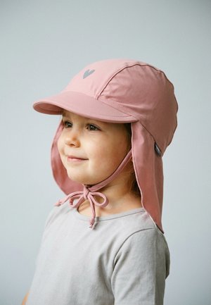 Young child wearing a light pink sun hat with neck flap and chin tie, looking to the side and smiling slightly against a plain background.