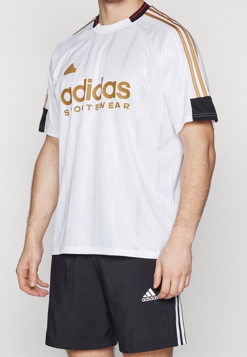 Vit Adidas sporttröja med guldlogga, svarta accenter på ärmarna och subtila vertikala ränder; ihop med svarta shorts med vit logga.