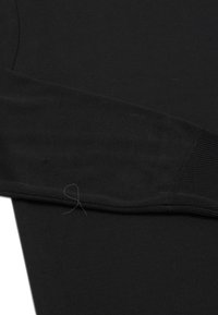 Maglietta nera a manica lunga con texture a coste. Presenta un design aderente e polsini elasticizzati. Leggermente visibile un filo sfilacciato vicino alla cucitura.