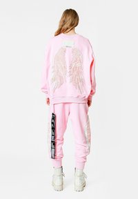 Conjunto de sudadera y pantalones rosa que presenta diseños metálicos de alas en la parte posterior, puños elásticos y una etiqueta de parche. El material parece suave y texturizado.