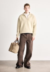 Béžový vlnený sveter s výstrihom do V a širokými manžetami, v kombinácii s tmavohnedými širokými nohavicami. Drží béžovú tašku s hnedými akcentmi.