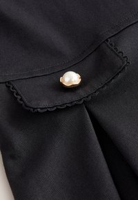 Tessuto nero con bordo smerlato e un bottone dorato con una grande perla bianca al centro.