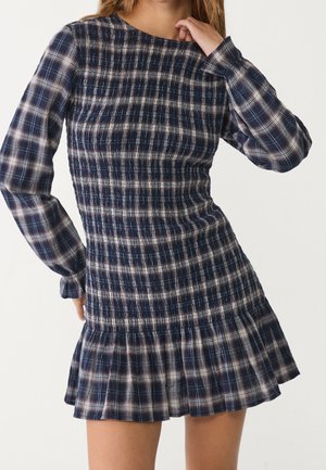 Femme portant une robe ajustée à carreaux bleu marine avec des manches longues et un ourlet à volants, main posée près du cou, sur un fond uni.