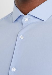 Jasnoniebieska koszula z odstającym kołnierzykiem i zapiętymi trzema białymi guzikami, noszona przez osobę o brązowej karnacji.