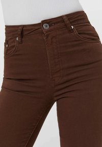 Vaqueros ajustados marrones con una tela texturizada, de tiro alto y con bolsillos frontales. Presenta detalles de botón metálico y cremallera.