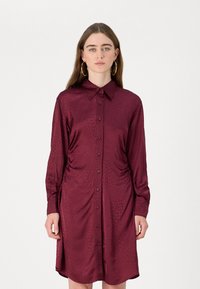 Vestido botão em marrom, feito de um tecido suave e leve. Apresenta um colarinho, lados franzidos e mangas longas com punhos de botão.