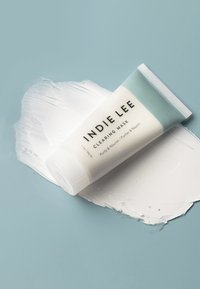Tube de compression blanc avec un capuchon bleu clair étiqueté "INDIE LEE CLEARING MASK", reposant sur une éclaboussure de crème blanche sur un fond bleu clair.