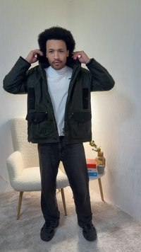 Άνδρας που ρυθμίζει τη κουκούλα του σκουρό πράσινου μπουφάν, φορώντας λευκό μπλουζάκι, μαύρο τζιν και μαύρα παπούτσια, στέκεται μέσα σε εσωτερικό χώρο κοντά σε λευκή πολυθρόνα και μικρό τραπέζι.