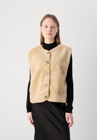 Beige fleeceväst med rund halsringning, framknäppning och något oversized design, lager på lager över en svart långärmad topp.