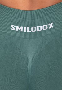 Leggings sportivi verdi con una vita a coste che presenta la scritta bianca "SMILODOX." Il tessuto appare liscio e aderente.