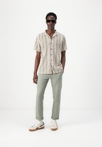Chemise à manches courtes rayée beige et blanche avec des boutons noirs, associée à un pantalon vert et des baskets blanches avec des semelles brunes.