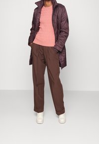 Veste matelassée violette, pull en maille torsadée rose, pantalon plissé marron et chaussures couleur crème. Tenue décontractée en couches avec textures variées.