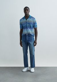 Kék hullám mintás rövid ujjú galléros ing, gallérral és gombokkal, egyenes szárú kék farmerral és fehér tornacipővel párosítva.