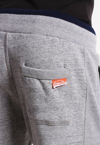 Graue Jogginghose aus weichem Stoff, mit geripptem Bund in Marineblau, Seitentasche und einem kleinen roten SuperDry-Logo-Patch.