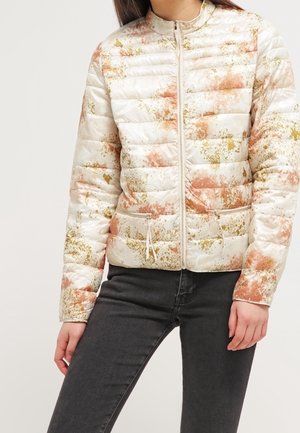 Femme portant une doudoune blanche zippée avec des motifs éclaboussés orange et or, associée à un jean noir, debout avec une main le long du corps.