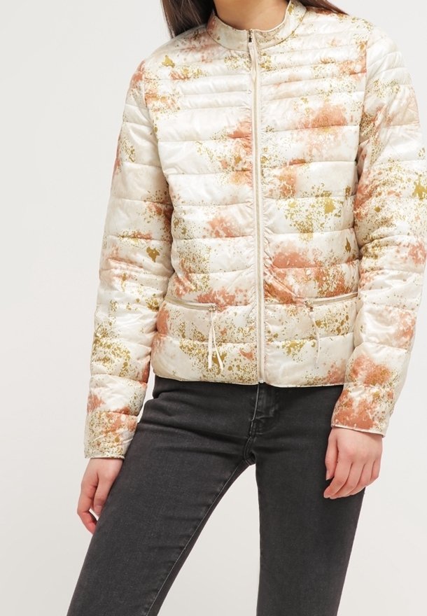 Femme portant une doudoune blanche zippée avec des motifs éclaboussés orange et or, associée à un jean noir, debout avec une main le long du corps.