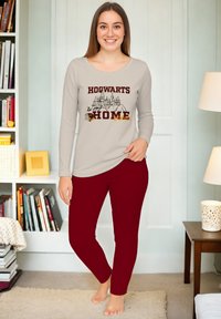 Donna sorridente in piedi a piedi nudi in una stanza accogliente, con una maglietta beige a maniche lunghe con la scritta "Hogwarts è casa mia" e pantaloni borgogna.