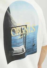 Hvid t-shirt med en stor grafisk print af en kystscene med en sejlbåd, tekst der lyder "ORGNLS," og en blød farvepalette.