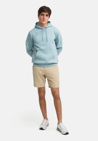 Felpa leggera blu con cappuccio e cordini regolabili, tasca frontale, abbinata a pantaloni corti beige e sneakers grigie; outfit casual, design minimalista.